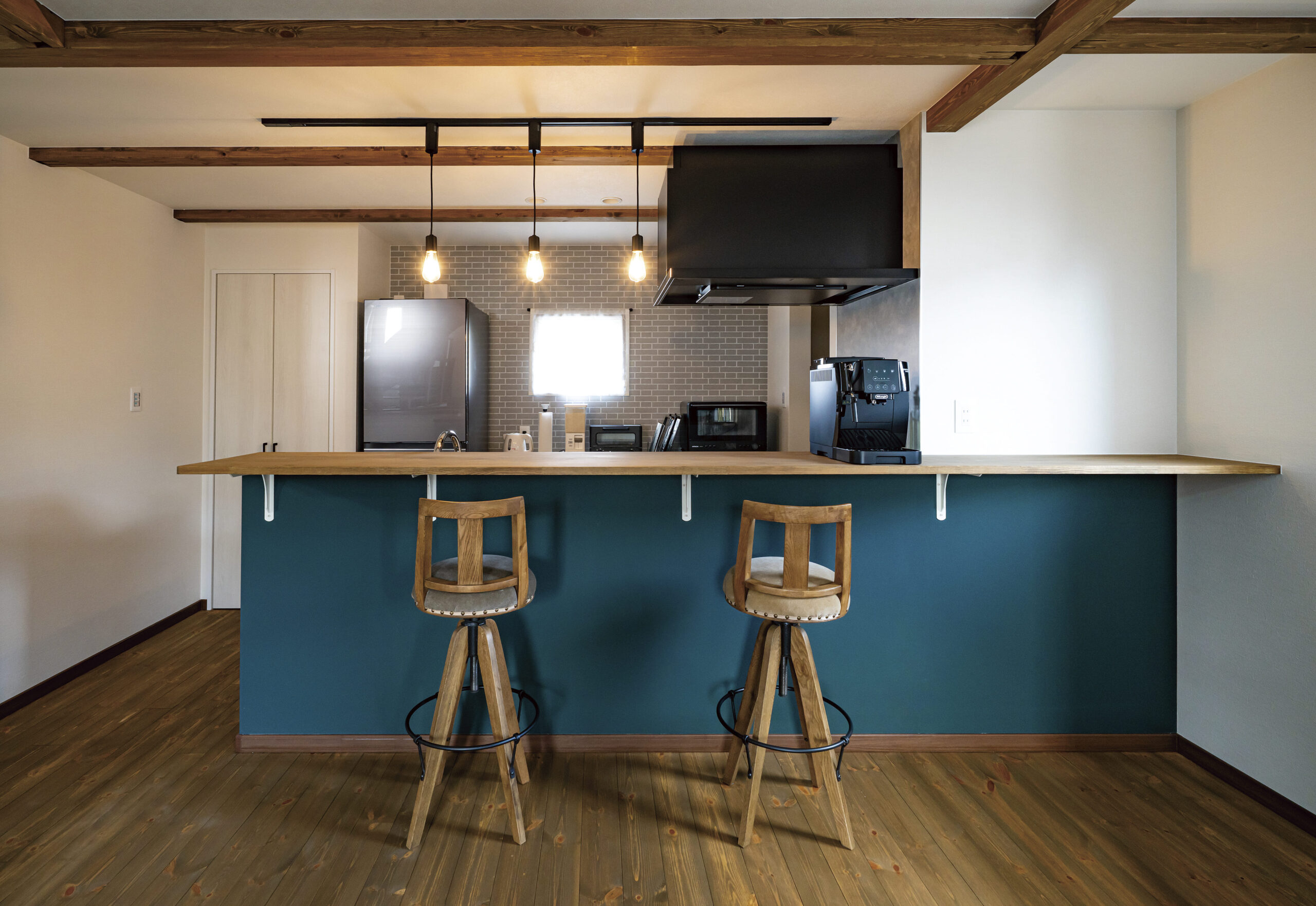 キッチンのカウンター下には外壁カラーに合わせたアクセントクロスを。シックな色味の床や梁とも調和して絵になる空間に。生活感をなるべく出さないよう、キッチンの奥にパントリー、冷蔵庫の横に扉付き収納を設け、食料品や生活用品を収納。室内がすっきりと片付くのが嬉しい。