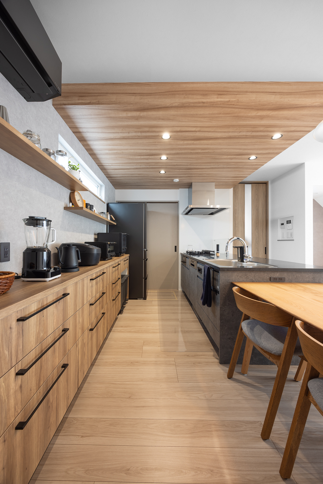グレーのフラットキッチンとブラック家電が映える空間。木目収納との調和で上質な雰囲気に仕上がった。