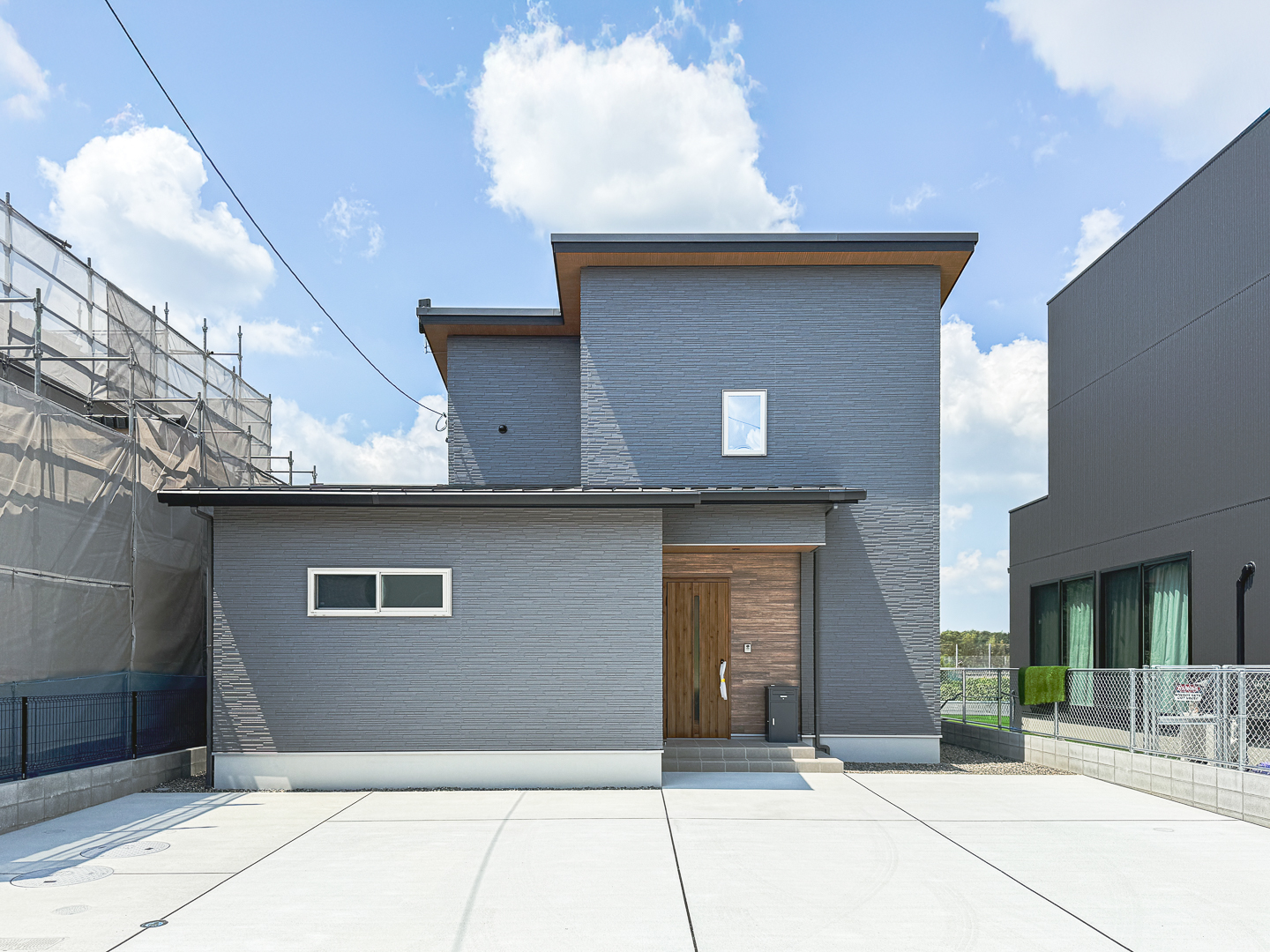 落ち着いたグレーの外壁に木目の玄関ドアが映えるシンプルな表情。道路側は控えめに、住まいは奥の庭と緑へ気持ちよくひらく構え。駐車スペースもゆったり。
