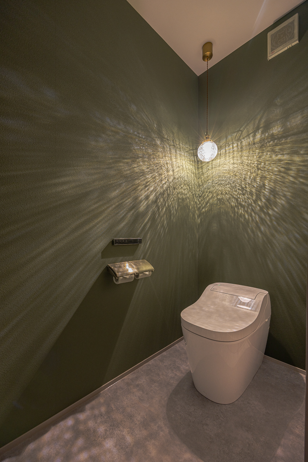 深い緑色のクロスが印象的なトイレ。天井から吊るされたペンダントライトが繊細な光の模様を壁面に投影し、空間に奥行きと劇的な表情を与えている。
