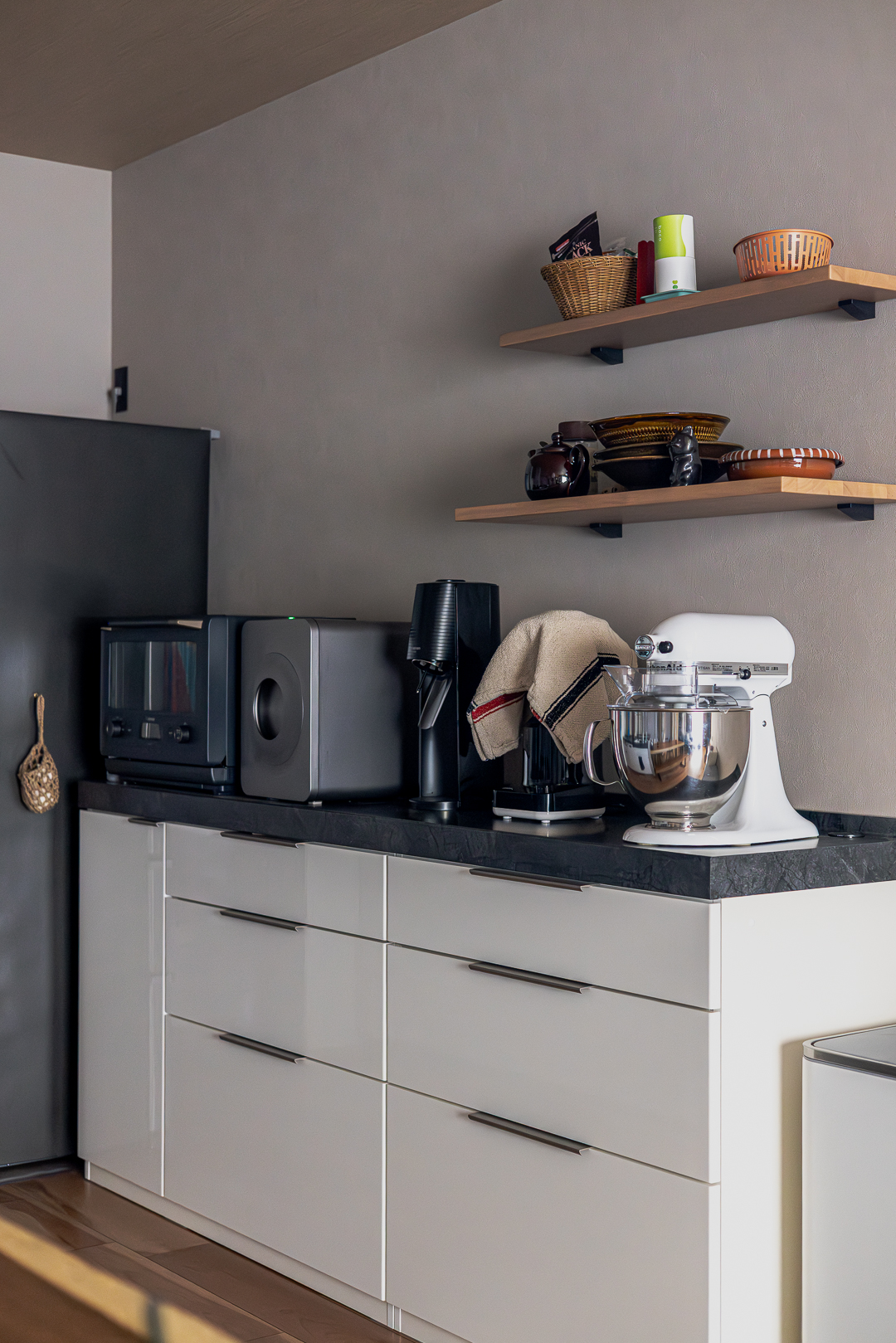 白いキャビネットに黒の天板を組み合わせたカップボード。オープン棚には食器類が並び、カウンターにはこだわりの調理家電が並ぶ機能的な空間だ。